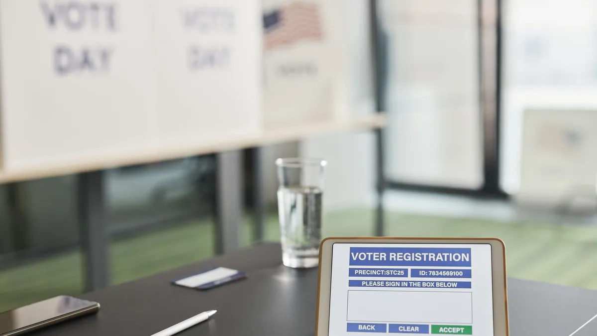 A tablet displaying a voter registration form on a table with a pen, phone, and glass of water, set against a backdrop of voting signs and a